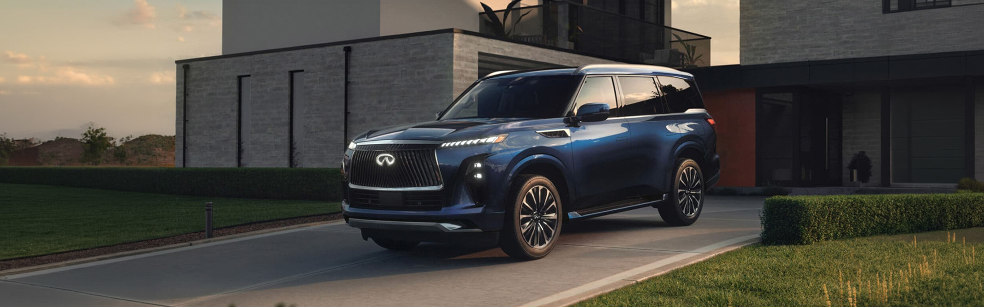 A blue 2026 INFINITI QX80 SUV parked in a modern driveway next to a stylish house at sunset.